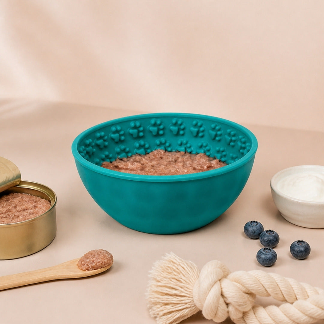 Bol de léchage turquoise garni de pâtée, avec boîte de pâtée, spatule en bois, myrtilles, yaourt nature et jouet en corde.